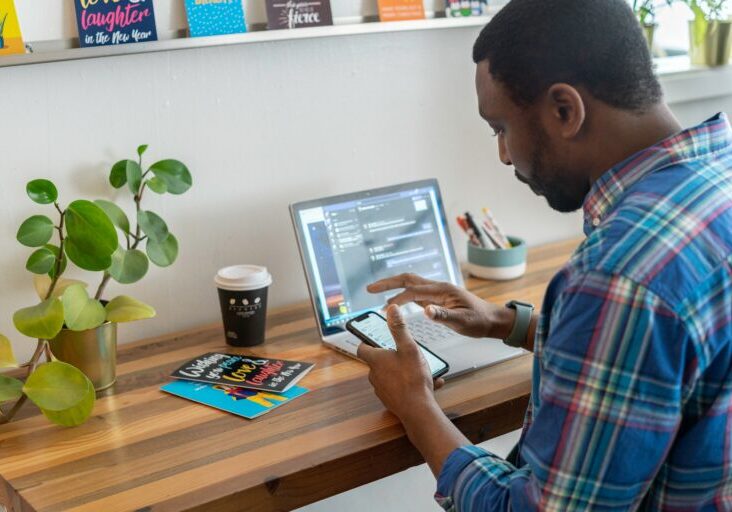 Stock image of a person of color using their phone and laptop at a desk.